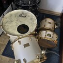 Bílá bicí souprava s pozlaceným hardwarem. Na basovém bubnu je nápis Pearl, na ostatních bubnech jsou štítky s nápisy Pearl a Export Series. V pozadí…