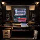 Fotografie zachycuje kompletní studio s pracovním stolem z tmavého dřeva. Na stole jsou dva počítačové monitory s otevřeným softwarem pro nahrávání,…