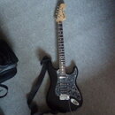Černá elektrická kytara s tvarem typu Stratocaster a hmatníkem z tmavého dřeva. Na hlavě je viditelné logo "Squier". Kytara má stříbrný pickguard s t…