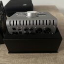 Stříbrné audio rozhraní v kompaktním kovovém provedení s žebrovaným horním krytem a velkým ovládacím knoflíkem. Na zadním panelu jsou vidět nápisy LI…
