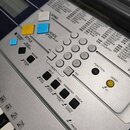 Detailní pohled na ovládací panel stříbrného elektronického klavíru značky YAMAHA. Jsou vidět různé ovládací tlačítka v barvách bílé, žluté a modré,…