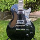 Černá elektrická kytara s tvarem singlecut umístěná na stojanu v trávě. Nástroj má dva humbuckery s průhlednými kryty a čtyři ovládací knofíky. Na ho…