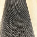 Fotografie zachycuje detail černé textilie s hrubou pletenou strukturou. Materiál je srolovaný do válce a položen na světlém dřevěném povrchu. Na sní…