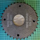 Černý kulatý součástka s ozubeným okrajem a čtyřmi otvory. Na povrchu je dvakrát uveden nápis USE M6 Metric Bolts. Uprostřed se nachází kruhový kovov…