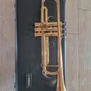 Zlatý mosazný kornét položený na černém tvrdém kufru. Na kufru je viditelné bílé logo a nápis YAMAHA. Nástroj vykazuje mírné známky opotřebení povrch…
