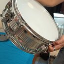Fotografie zachycuje snare drum s chromovaným kovovým korpusem a bílým membránou. Nástroj drží osoba v rukou. Na viditelných částech nástroje nejsou…