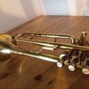 Mosazná trumpeta s patronskými ventily a bílými krytkami. Nástroj vykazuje známky opotřebení povrchu, jsou vidět drobné škrábance a oxidace. Žádné zn…