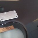 Detail korpusu elektrické kytary v tmavě hnědé barvě s vysokým leskem. Na zadní straně je vidět kovová krytka s nápisem Ibanez, připevněná čtyřmi šro…