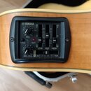 Detail ovládacího panelu akustické kytary značky YAMAHA. Černý plastový panel obsahuje dva otočné ovladače s nápisy VOL a MIC. VOL, přepínač PHASE a…
