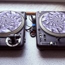 Dvě černé gramofony s 탈-slipmaty s abstraktním fialovo-bílým motivem. Na levém slipmatu je čitelný nápis „almantronics“. Oba přístroje mají stříbrné…