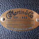 Oválný mosazný štítek připevněný na černý strukturovaný povrch pouzdra. Na štítku je nápis Martin & Co. a pod ním EST. 1833. Ve spodní části je text…