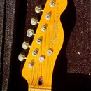 Fotografie zachycuje hlavu žluté elektrické kytary z dřeva. Na povrchu je černý nápis „Fender“ a vedle něj svisle orientovaný text „TELECASTER®“. Nás…