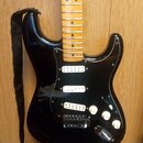Černá elektrická kytara s tvarem typu Stratocaster a černým pickguardem. Má světle hnědý javorový hmatník a tři bílé snímače. Na pickguardu je vidite…