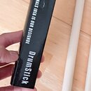 Černá papírová krabice s bílými nápisy „PROFESSIONALS IDEA IS OUR DESIGNDE“ a „DrumStick“. Vedle krabice leží dvě světlé dřevěné paličky na buben. Po…