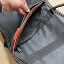 Černý textilní obal na hudební nástroj s oranžovými detaily u zipu. Na vnitřní straně kapsy je viditelná bílá štítková vložka s oranžovým logem a náp…