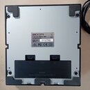 Fotografie zachycuje spodní stranu stříbrného elektronického zařízení s černými gumovými nožičkami v rozích. Uprostřed je nalepený štítek s logem ZOO…