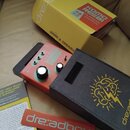 Na fotografii je efekt pedál červené barvy s nápisy "DREADBOX" a "KOMO REBI". Pedál má tři ovládací knoflíky, tři přepínače a je umístěn v šedém text…