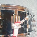 Detailní pohled na chromovaný mechanismus napnutí snareho na černém bubnu. Na kovovém dílu je svisle vyražen nápis SONOR. Mechanismus je fixován červ…