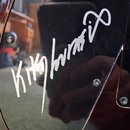 Detail černého, lesklého povrchu hudebního nástroje. Na povrchu je bílý nápis v podobě podpisu „Kiko Loures“. Jsou vidět dva kovové šrouby a odrazy o…