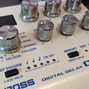 Bílý efekt pedál s nápisy BOSS, DIGITAL DELAY a DD-200. Na horní ploše jsou umístěny stříbrné kovové ovraty a černý displej. Jsou zde čitelné názvy f…