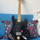 Černá elektrická kytara s mírně prohnutým tělem a světle hnědým hnedem hmatníkem. Na hlavě je nápis Squier Stratocaster. Tělo má černý pickguard, tři…