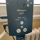 Černý ovládací modul elektronických bicích s kruhovým displejem. Na přední straně jsou bílá loga Roland a V-Drums, nápisy Tempo, Coach, Drums, Song a…
