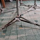 Fotografie zachycuje spodní část kovových stativů, pravděpodobně činelových nebo mikrofonních. Povrch kovových trubek a nožiček vykazuje výrazné opot…