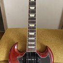 Elektrická kytara s červeným dřevěným korpusem a černým pickguardem. Nástroj má dva humbuckery a hnedlový hmatník s trapezoidními vložkami. Povrch ko…