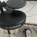 Černý polstrovaný bubeníckýstolek s nápisem YAMAHA a doprovodným logem. Povrch sedáku vykazuje mírné vrásnění materiálu. V pozadí je vidět část elekt…