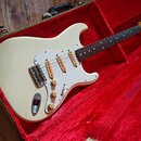 Bílá elektrická kytara typu Stratocaster s bílým pickguardem a hnedým hmatníkem, umístěná v otevřeném kufru s červeným polstrováním. Na hlavě hmatník…