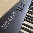Detailní pohled na ovládací panel a klávesy černého syntetizátoru. Na povrchu je bílý nápis Roland JV-30 a pod ním text 16 PART MULTI TIMBRAL SYNTHES…