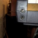 Detailní pohled na část černého plastového zařízení. Jsou zde vidět dva kulaté konektory a nad horním z nich je bílý nápis PHONES. Povrch plastu vyka…