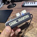 Černý efekt pedál s bílým nápisem CARL MARTIN. Na horní straně jsou dva bílé zapínací spínače s modrým podsvícením a jeden kovový přepínač. Zařízení…