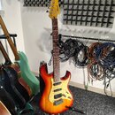 Elektrická kytara s tělem v červeno-žlutém sunburst provedení a bílým pickguardem s perleťovým vzorem. Na hlavě hmatníku je viditelné logo Cort. Kyta…