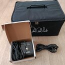 Černý textilní pouzdro s bílým nápisem Paul Reed Smith®. Vedle něj je otevřená kartonová krabice, ve které se nachází černý napájecí adaptér s nápise…