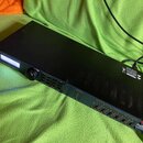 Černý rackový zesilovač s kovovým šasi a ventilačními otvory na bočních stranách. Na předním panelu je bíle podsvícený displej, ovládací knoflík a řa…