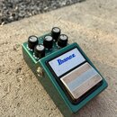 Zelený efekt pedál v kovovém šasi s pěti černými ovládacími knoflíky. Na bílém štítku je modré logo Ibanez a nápisy TS9B Bass Tube Screamer. Pod knof…