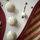 Detail elektrické kytary s bílým pickguardem a vínovým tělem. Na bílém plastovém krytu jsou umístěny tři bílé oválné potenciometry s číslováním 1–10…