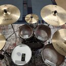 Celá bicí souprava s činely na vzorovaném koberci. Na činelech je vidět nápis „Istanbul Agop“, na bubnech značka „EVANS“ a na bílém snare bubnu nápis…