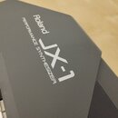 Detailní pohled na šedý plastový povrch syntetizátoru. Na povrchu je bílý nápis Roland a pod ním modelové označení JX-1 s doplňujícím textem PERFORMA…