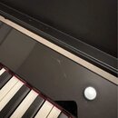 Detailní pohled na černý, lesklý povrch klávesového nástroje s bílými a černými klávesami. Na horní části černého panelu je viditelný škrábanec a mal…