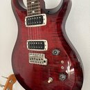 Elektrická kytara s červeným lakovaným korpusem z plamenového javoru a dvěma humbuckery s bílými rámečky. Na hmatníku jsou viditelné vkládky ve tvaru…