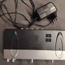 Černý a stříbrný elektronický přístroj s logem BEHRINGER v trojúhelníku a nápisem www.behringer.com. Zařízení má na horní straně čtyři konektory a v…