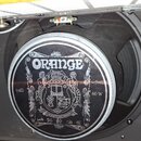 Fotografie zachycuje reproduktor v černém rámu, umístěný uvnitř skříně. Na středovém krytu je bílý nápis ORANGE a dekorativní grafika. Po stranách js…