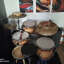 Celá bicí souprava s přirozeným dřevěným povrchem v odstínech žluté a hnědé. Na bubních je viditelné logo "Mapex", na jednom z činelů nápis "B MEINL"…