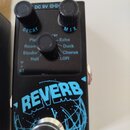 Černý efekt pedál s nápisem REVERB v modré barvě a grafickým motivem sítě. Na horní části jsou tři černé oválné knoflíky s popisky DECAY, MIX a sezna…