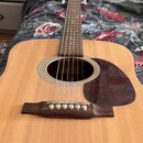 Akustická kytara s přírodním dřevem na desce a hnědým hmatníkem. Na desce je tmřečervený pickguard s mramorovým vzorem. Nástroj nemá žádné viditelné…
