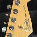 Fotografie zachycuje hlavu elektrické kytary z přírodního světlého dřeva s chromovanými ladičkami. Na povrchu je černý nápis Fender® STRATOCASTER a n…