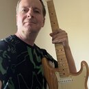 Fotografie zobrazuje muže s elektrickou kytarou typu Stratocaster. Kytara má přírodní dřevěnou barvu korpusu, světlý hmatník a krémový pickguard. Na…