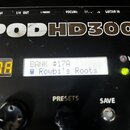 Detailní pohled na ovládací panel černého efektového procesoru s velkým bílým nápisem PODHD300. Na displeji je text BANK #17A a Rowbi's Roots, vedle…